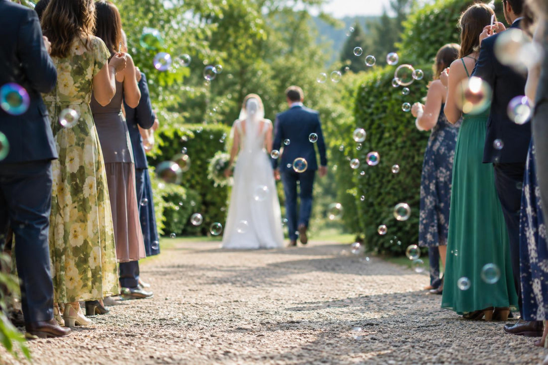 Wedding guests line a garden path as bubbles float around a couple walking away in daylight
