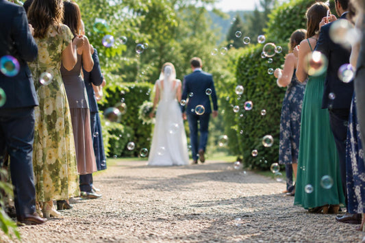 Wedding guests line a garden path as bubbles float around a couple walking away in daylight